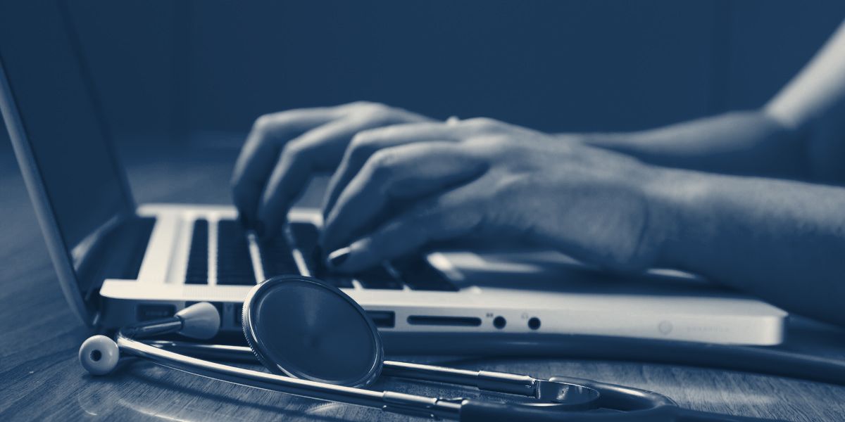 Mãos digitando no notebook ao lado de um estetoscópio